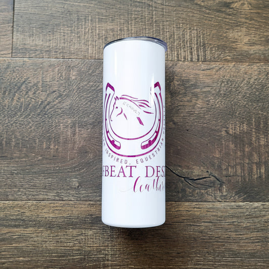 A white tumbler with a purple logo of Hoofbeat Designs, placed on a wooden surface.
