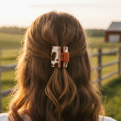 Person with long brown hair wearing a cow print hair clip, standing in a field with a wooden fence and barn in the background.